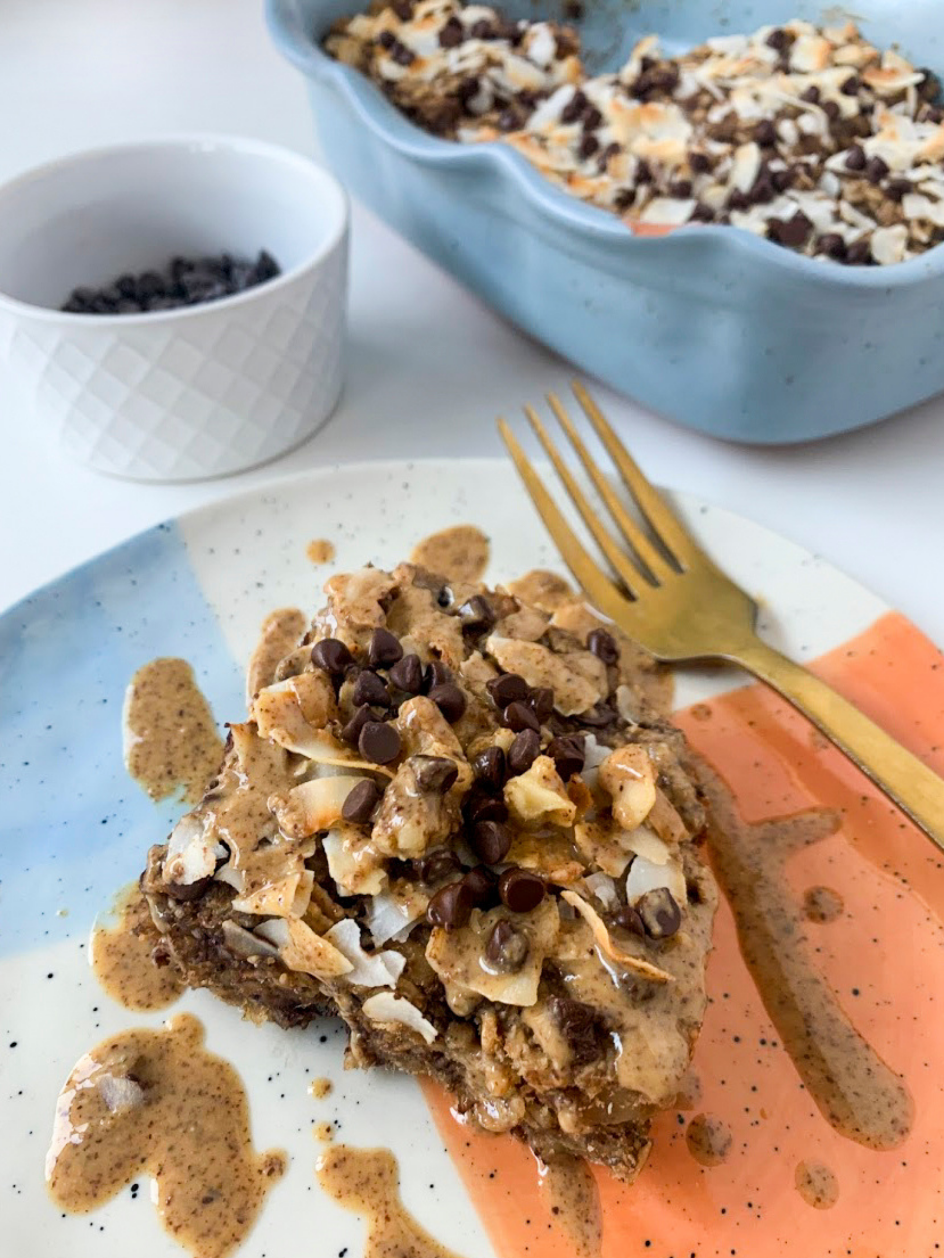 baked oatmeal with chocolate chips, coconut flakes, and almond butter on a plate with a fork. Baking dish and bowl of chocolate chips in background.